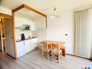 Cozinha compacta com armários brancos, micro-ondas e cafeteira. Há uma mesa de jantar de madeira com cadeiras.