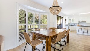 A modern kitchen with white cabinets, a wooden dining table, wicker chairs, and a large window with blinds. - Seagrass - Pet Friendly - 10 Mins Walk to Beach (Vincentia)