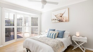 A bedroom with a bed, pillows, a nightstand, a lamp, a ceiling fan, and a large window with blinds. - Seagrass - Pet Friendly - 10 Mins Walk to Beach (Vincentia)