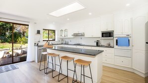 A modern kitchen with a central island, bar stools, white cabinetry, and a view of the outdoors. - Seagrass - Pet Friendly - 10 Mins Walk to Beach (Vincentia)