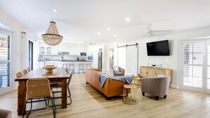 A spacious living area with a dining table, sofa, and chairs, a kitchen in the background, and a TV mounted on the wall. - Seagrass - Pet Friendly - 10 Mins Walk to Beach (Vincentia)