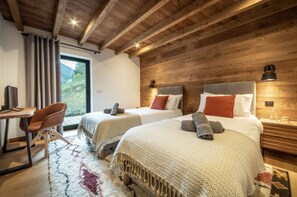 Quarto aconchegante com duas camas, vigas de madeira no teto, uma mesa de trabalho e vista para o verde lá fora.