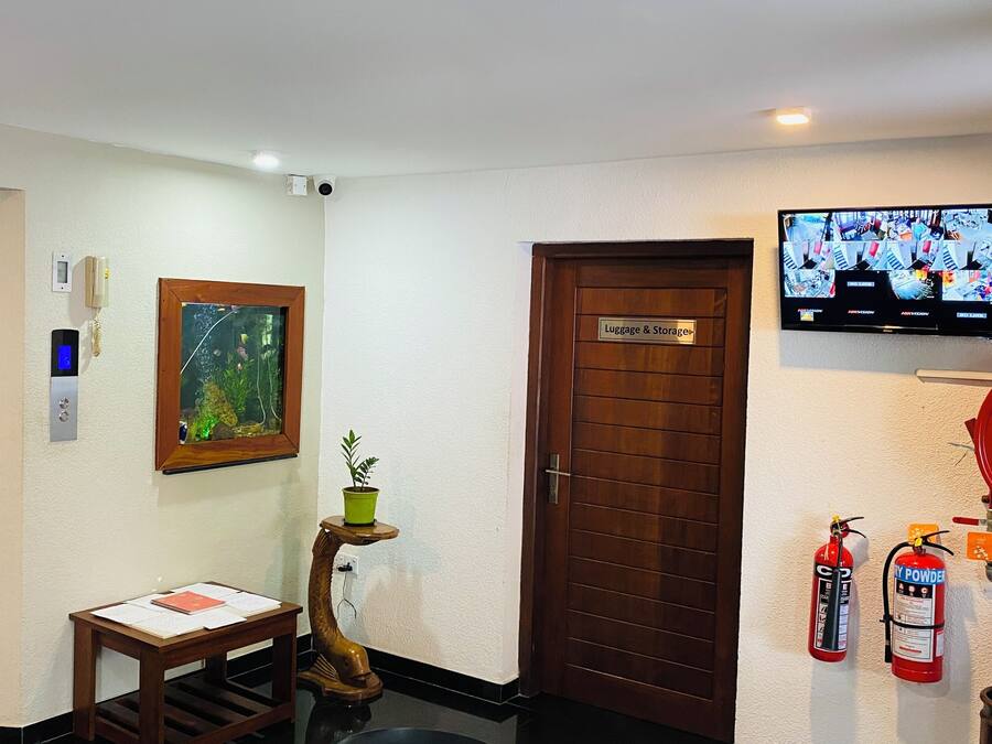 Een hotel lobby met een houten deur waarop 'Bagage & Opslag' staat, een wandtelevisie, een brandblusser, een kleine tafel met papieren en een potplant.