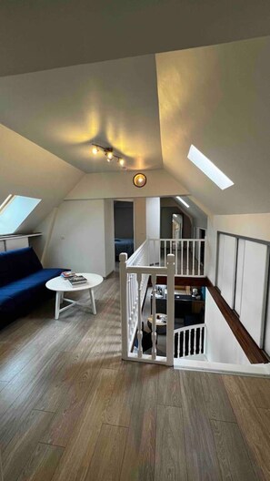 Habitación estilo loft con techo inclinado, escalera, sofá azul, una pequeña mesa redonda y barandilla blanca.