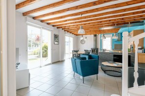 Amplia sala de estar con techo alto y vigas de madera a la vista, comedor con sillas azules y cocina con electrodomésticos modernos.