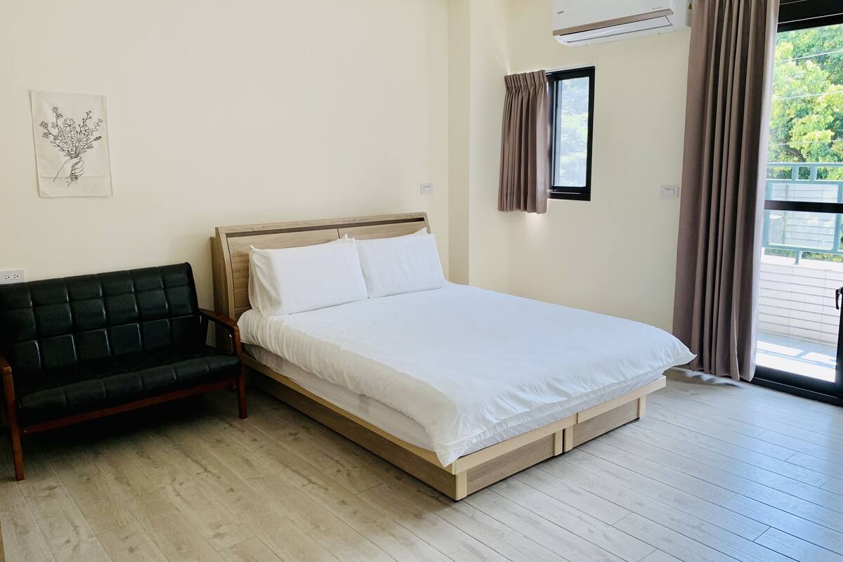A bedroom with a bed, a sofa, a window with curtains, and a wall-mounted air conditioner.