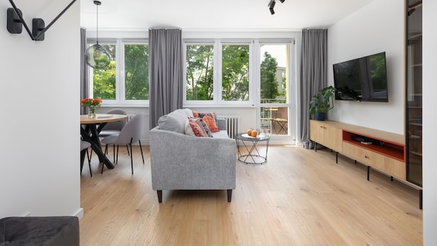 Una sala de estar moderna con un sofá gris, una mesa de comedor y sillas, un televisor en la pared y ventanas amplias con cortinas.