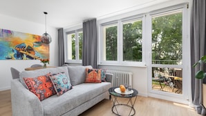 Una sala de estar moderna con un sofá gris, cojines coloridos, una mesa de vidrio con fruta y ventanas grandes que ofrecen vistas al exterior.