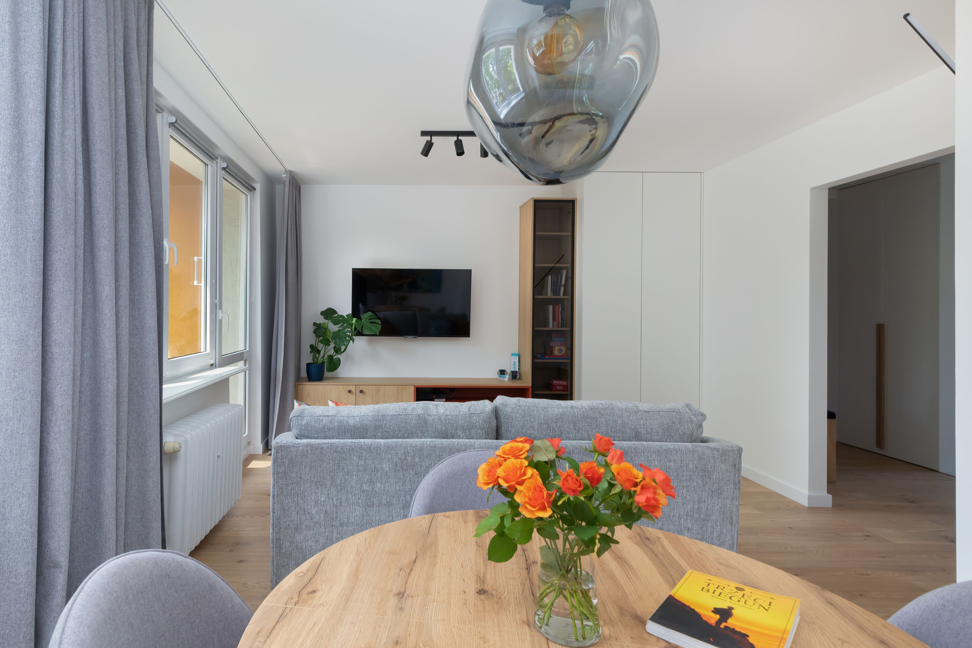 Een moderne woonkamer met een grijze bank, een houten salontafel met een boek en bloemen, een televisie aan de muur en een boekenkast.