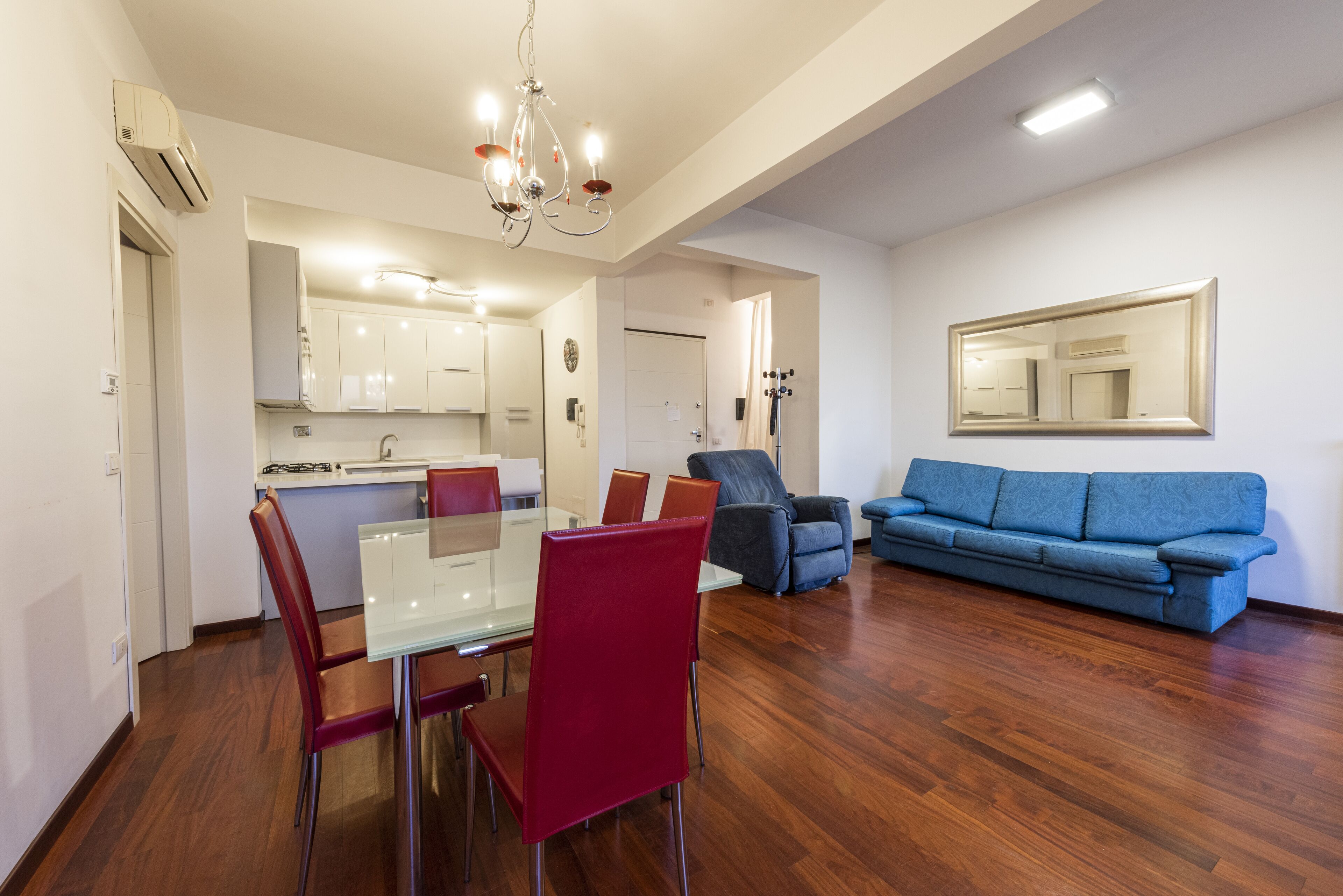 A modern living room with a glass dining table, red chairs, a blue sofa, and a chandelier.