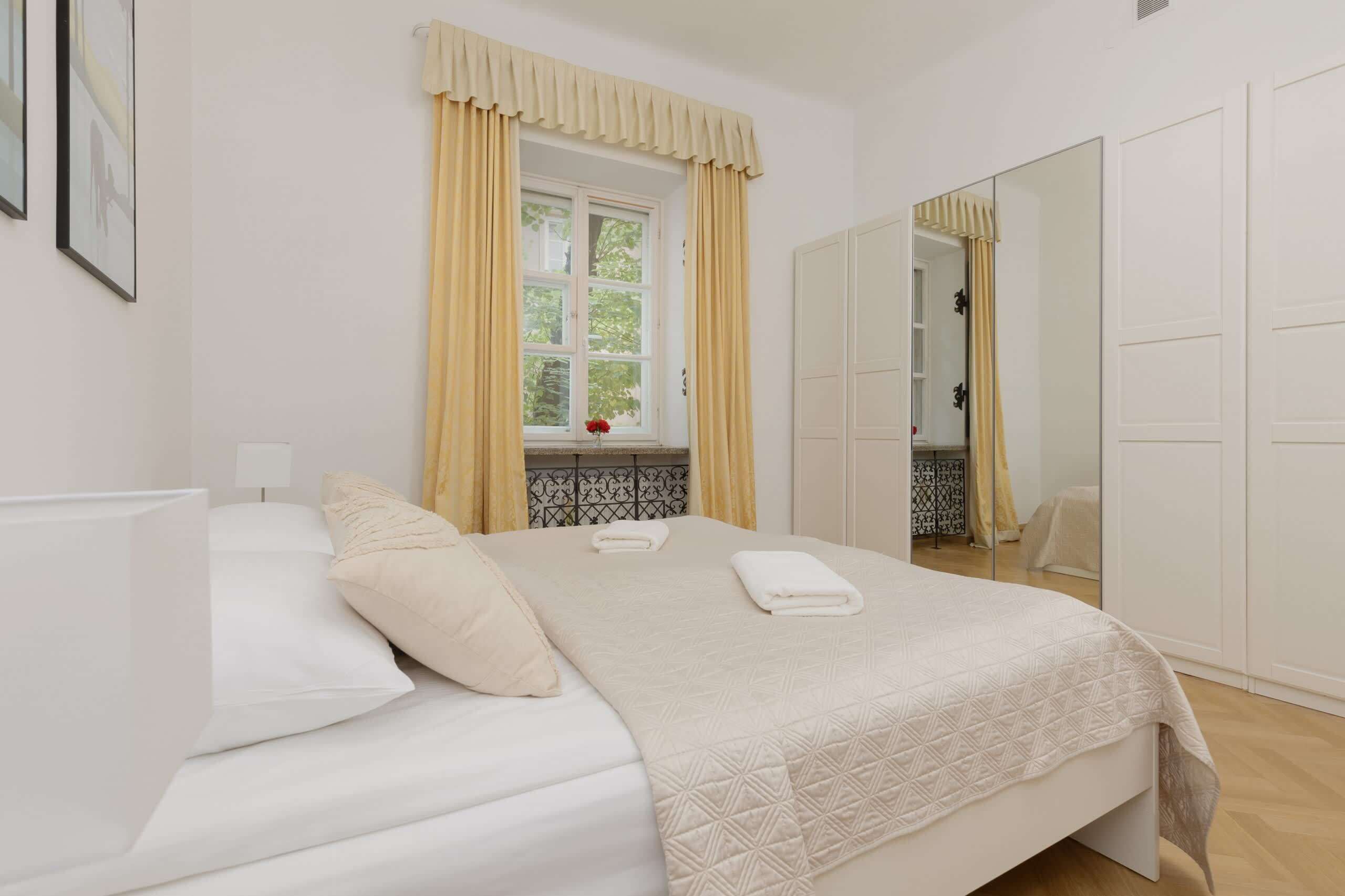 Un dormitorio ordenado con cama, ventana con cortinas, espejo y armario.