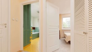 Una habitación de hotel con piso de madera, una cama con edredón blanco, una ventana con persianas y una puerta verde que da a un baño con piso amarillo.