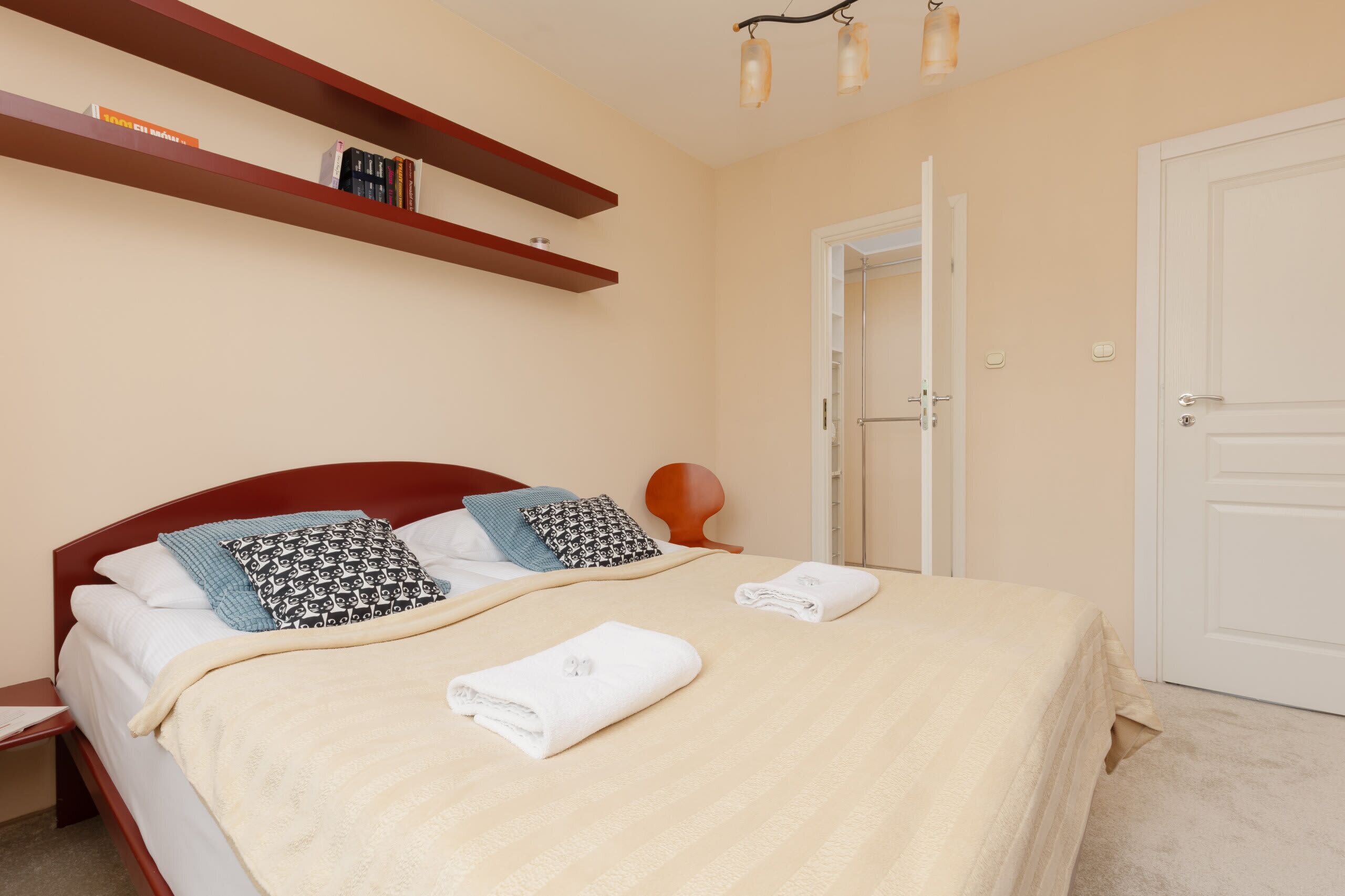 Un letto ben rifatto con una coperta beige, due cuscini e due asciugamani. Sopra il letto, due mensole in legno con libri e una piccola lampada.