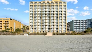 A high-rise hotel with multiple balconies, a sandy beach in the foreground, and a clear sky. - Oceanfront Efficiency T401 (Myrtle Beach)
