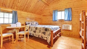 Ein gemütliches Blockhauskabinenzimmer mit Holzbalkendecke, einem Bett mit gemustertem Überwurf, einem kleinen Tisch mit Stühlen und einem Fenster mit Vorhängen.