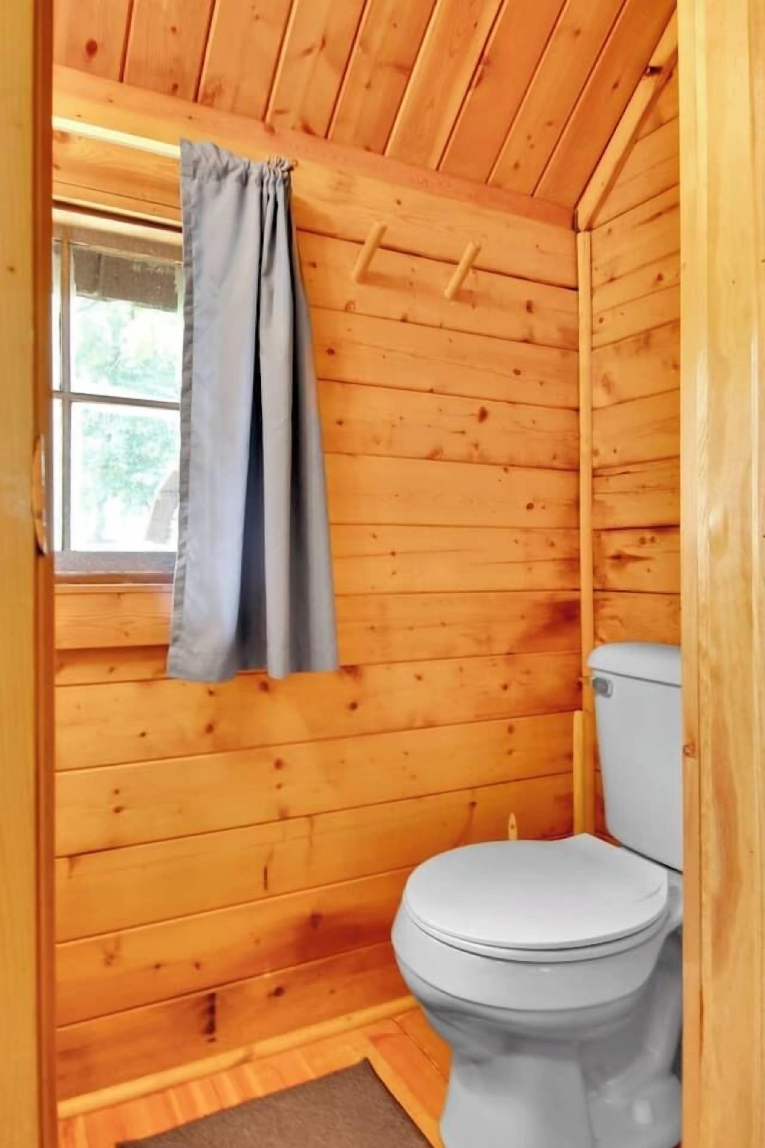 Een houten badkamer met een toilet, een raam met gordijn en een houten plafond.