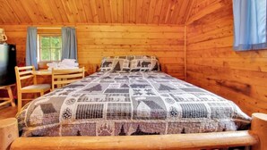 Habitació d'una cabana de fusta amb un llit, una taula, cadires i una finestra amb cortines.