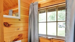 Bany d'una cabana de fusta amb un rentapans blanc, armaris de fusta, un mirall i una finestra amb cortines.