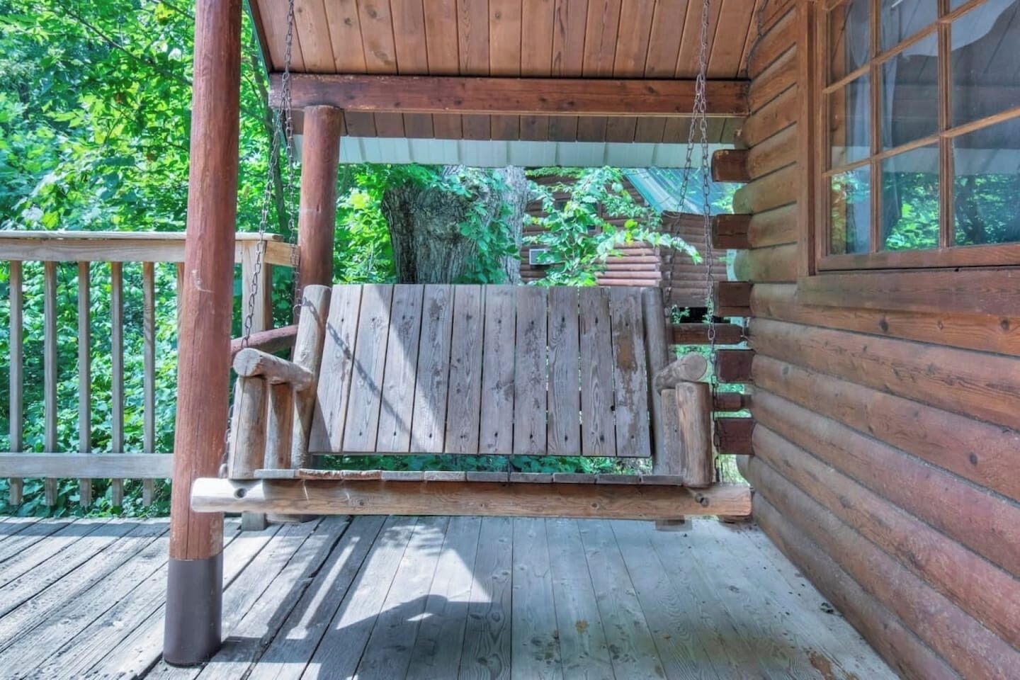 A wooden porch with a swing, a wooden railing, and a view of greenery.