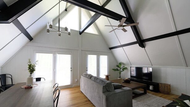 A spacious loft with a high ceiling, exposed beams, a grey sofa, a flat-screen TV, and a dining area with a table and chairs. - Stunning Aframe Cabin with Deck (Bloomington)