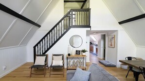A spacious loft with a high ceiling, a wooden staircase, a gray sofa, two wooden chairs, a small table with decorative items, and a view into the kitchen area. - Stunning Aframe Cabin with Deck (Bloomington)