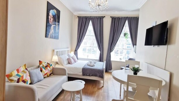 A modern bedroom with a bed, a sofa, a small table, a TV, and a window with curtains. - Lovely Renovated Large Spacious Georgian Studio (Dublin 1)