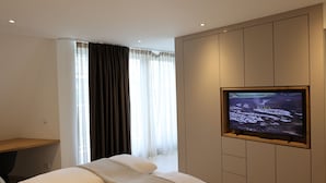 Ett modernt hotellrum med en stor, platt-TV som är monterad på väggen, en säng med ett vitt sänggavel, ett skrivbord i trä och ett fönster med tunna, genomskinliga gardiner.