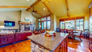 A spacious living area with a stone fireplace, wooden ceiling beams, and a large window offering a view of trees.