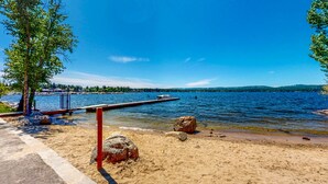 A sandy beach with a dock extending into a lake, surrounded by trees and a clear blue sky.