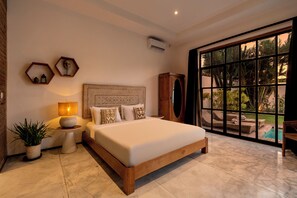 A bedroom with a large bed, a nightstand, a lamp, a wall-mounted mirror, and a view of a pool through a large window. - Villa Maje by Alfred in Bali (Seminyak)