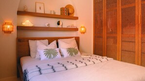 A neatly made bed with a patterned bedspread, two white pillows with palm leaf designs, and a wooden headboard. Above the bed, there are two wooden shelves with decorative items and two wall-mounted lamps. - Vacation apartment Matimar, Costa Calma, terrace, WiFi (Costa Calma)