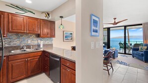 A kitchen with wooden cabinets, a dishwasher, and a backsplash with blue and white tiles. There is a dining area with a view of the ocean through large windows. - 1bd Oceanfront gem with charm, views & Island vibe (Lahaina)