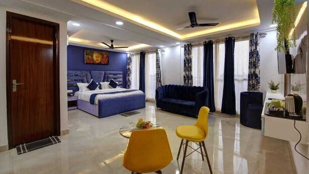 A modern hotel room with a bed, a sofa, a dining table with chairs, and a ceiling fan. - Hotel Noida Dreamz 144 (Noida)