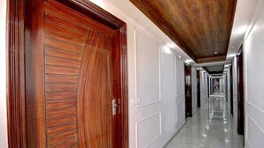 A long, narrow hallway with a wooden door on the left and white walls. - Hotel Noida Dreamz 144 (Noida)