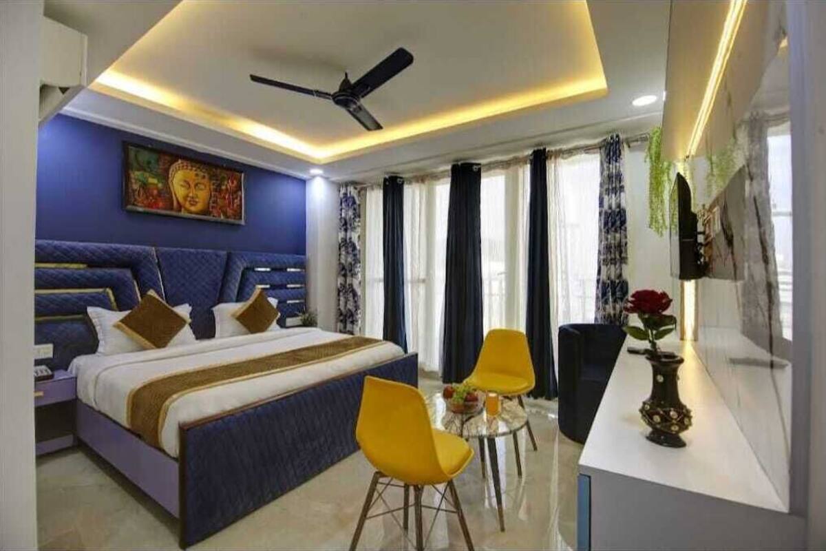 A modern hotel room with a large bed, a ceiling fan, a dining table with yellow chairs, and a vase with a red flower.