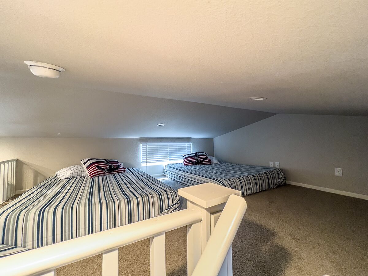 A small, single-level room with two beds, a white desk, and a window with blinds.