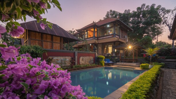 Una casa de dos pisos con piscina, rodeada de vegetación exuberante y flores moradas.