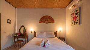Habitación de hotel con una cama grande, dos mesitas de noche, un aparador de madera, un espejo, un ventilador de pared y un cuadro con motivos florales.