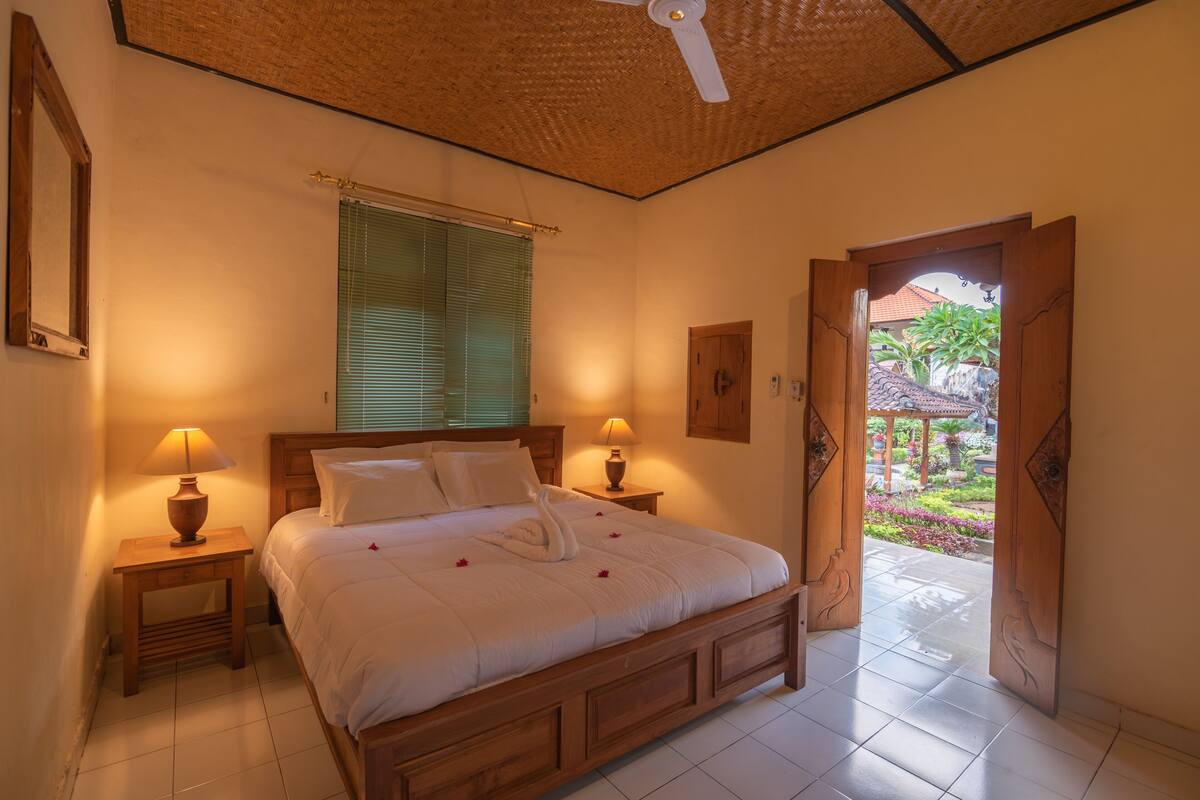 A bedroom with a wooden bed, two bedside lamps, a window with blinds, and a door leading to an outdoor area.