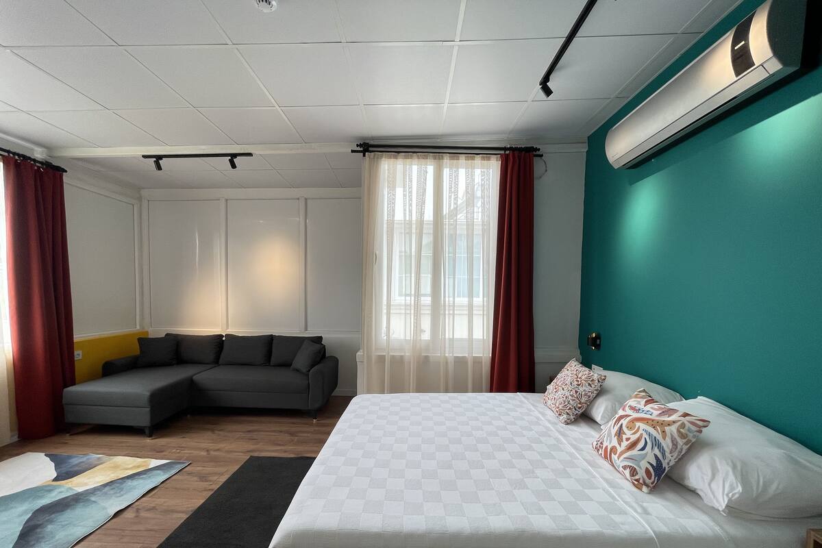 Quarto de hotel moderno com uma cama grande, um sofá cinzento, um pequeno espaço de estar e vista da cidade através de cortinas translúcidas.