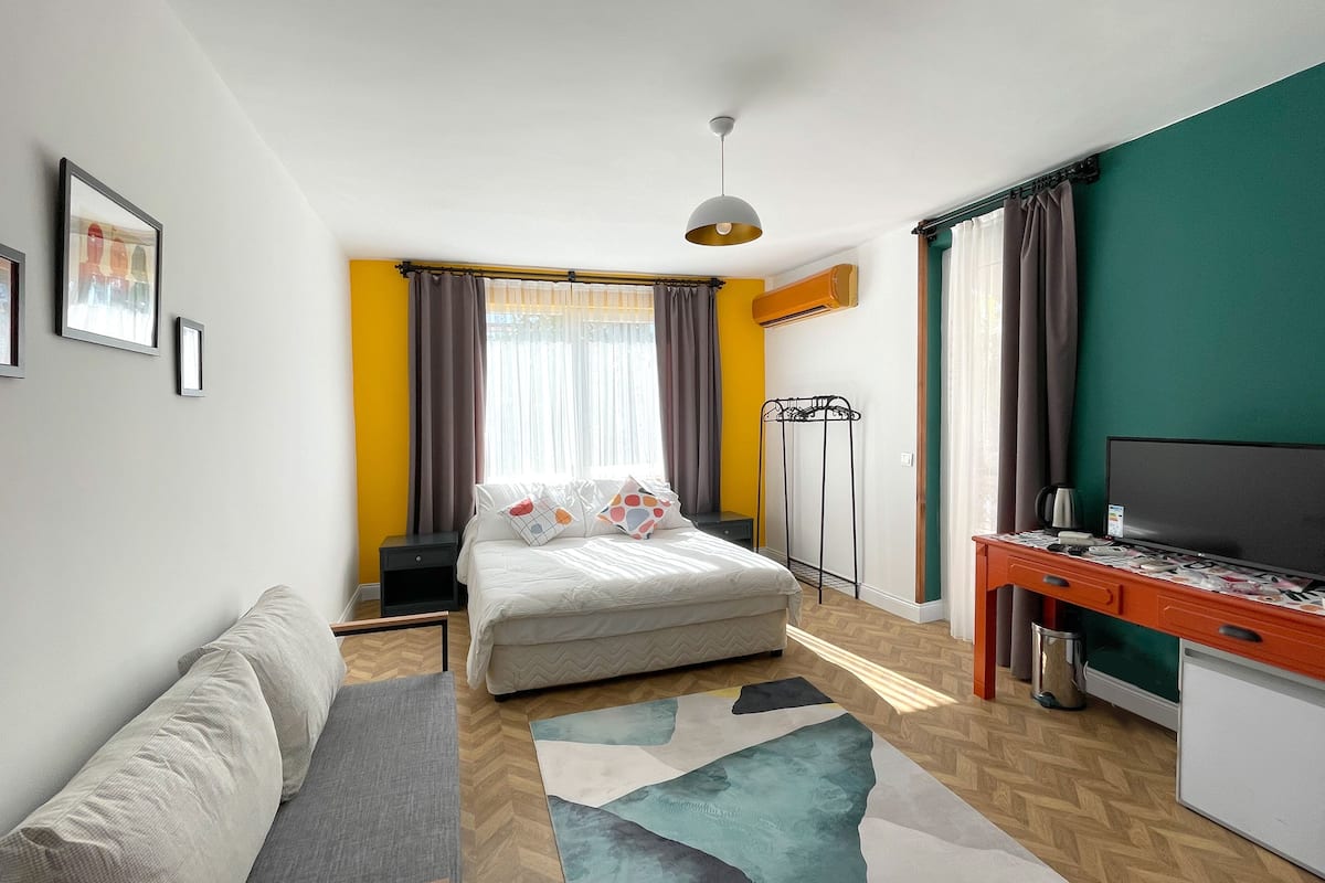 Quarto de hotel moderno com cama, escrivaninha, televisão, sofá e tapete estampado.