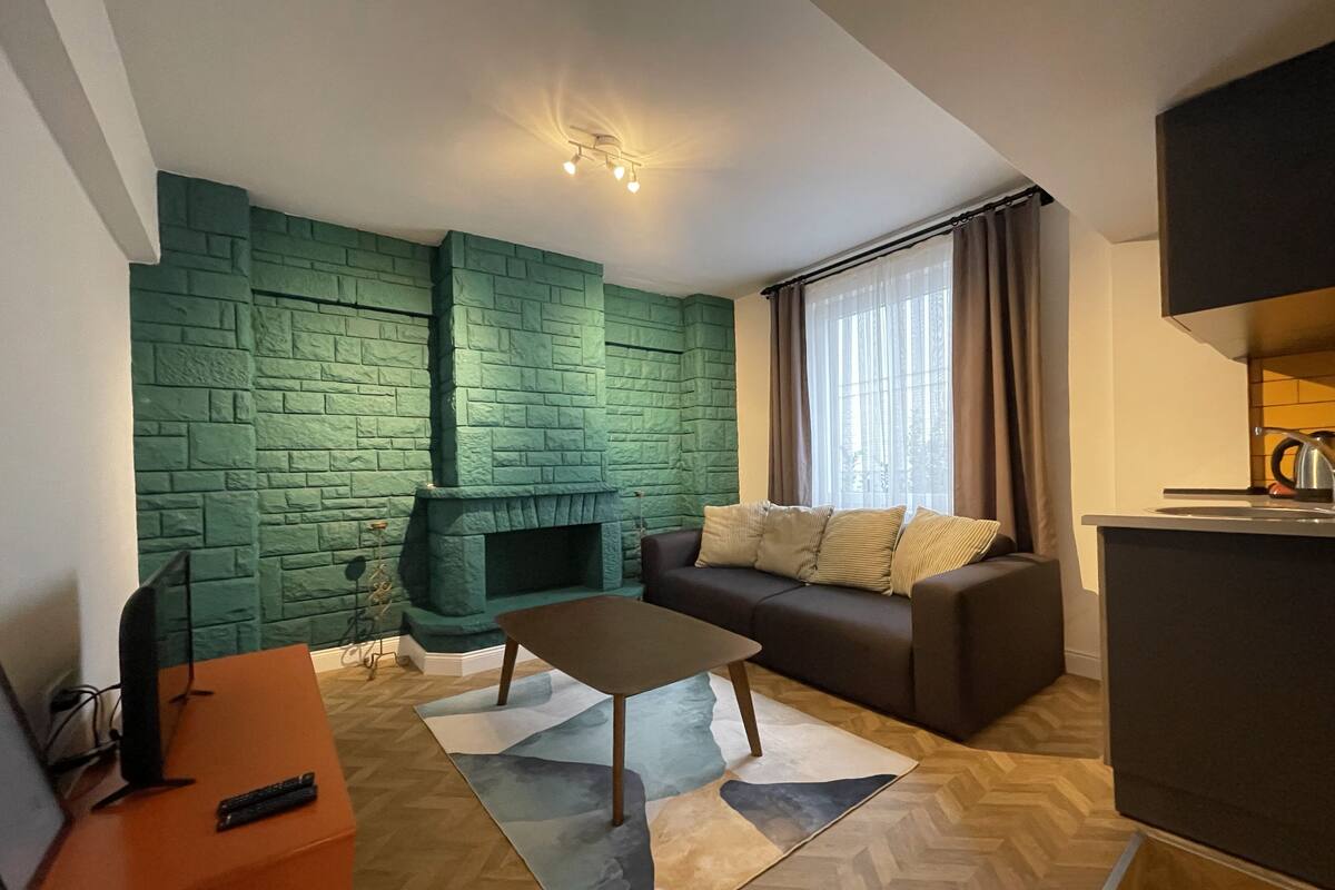 Sala de estar com parede de tijolos verdes, lareira, sofá, mesa de centro e área de cozinha com chaleira.
