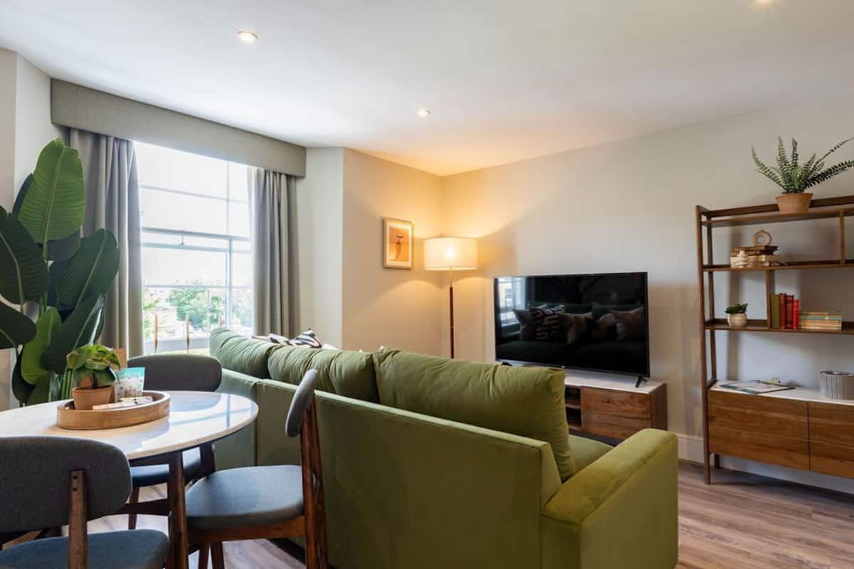 Een moderne woonkamer met een groene bank, een flatscreentelevisie, een eettafel met stoelen, een lamp en een boekenkast.
