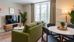Ein modernes Wohnzimmer mit einem grünen Sofa, einem Flachbildfernseher, einem Esstisch mit Stühlen und einer Topfpflanze.