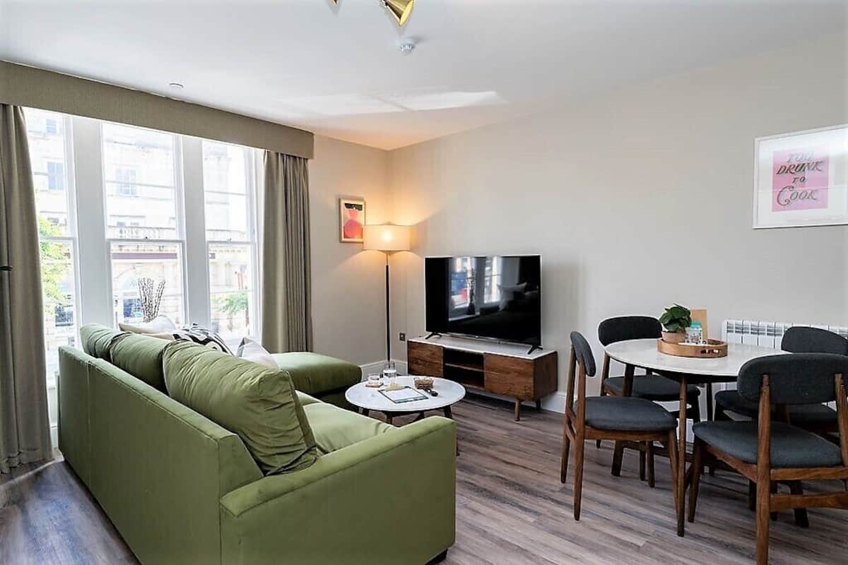 Een woonkamer met een groene bank, een televisie, een eettafel met stoelen en een raam met gordijnen.