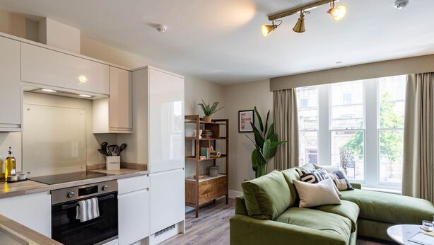 Eine moderne Küche mit wei ßen Schränken, einem eingebauten Backofen und einer Holz Arbeitsplatte. Im Wohnbereich befindet sich ein grünes Sofa und ein kleiner Tisch, wobei ein großes Fenster für natürliches Licht sorgt.
