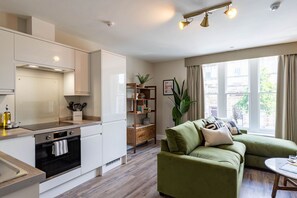 Une cuisine moderne avec des armoires blanches, un four encastré et un plan de travail en bois. Le salon est agrémenté d’un canapé vert et d’une petite table, et une grande fenêtre laisse entrer la lumière naturelle.