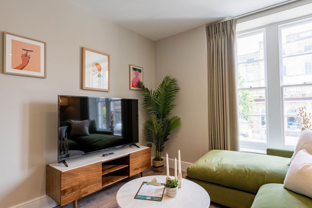 Un salon avec un téléviseur à écran plat posé sur un meuble en bois, un canapé vert, une table basse ronde ornée d’une plante et une fenêtre avec des rideaux.
