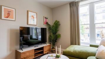 Un salon avec un téléviseur à écran plat posé sur un meuble en bois, un canapé vert, une table basse ronde ornée d’une plante et une fenêtre avec des rideaux.