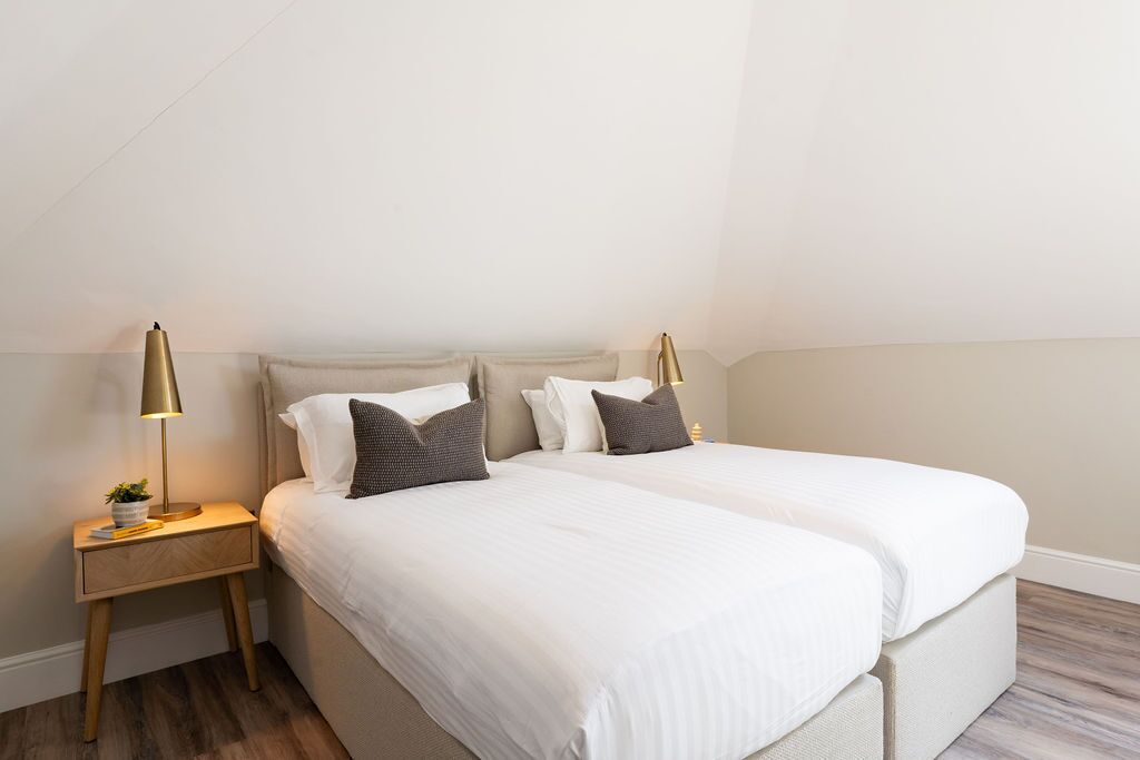 Ein Doppelbett mit weißer Bettwäsche und zwei Nachttischlampen, ein kleiner Nachttisch mit einer Pflanze und ein Holzboden.